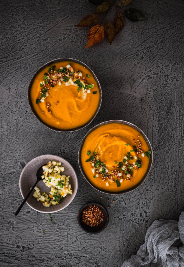 Zwei Schüsseln cremige Kürbissuppe in herbstlichem Setting auf grauem Untergrund.