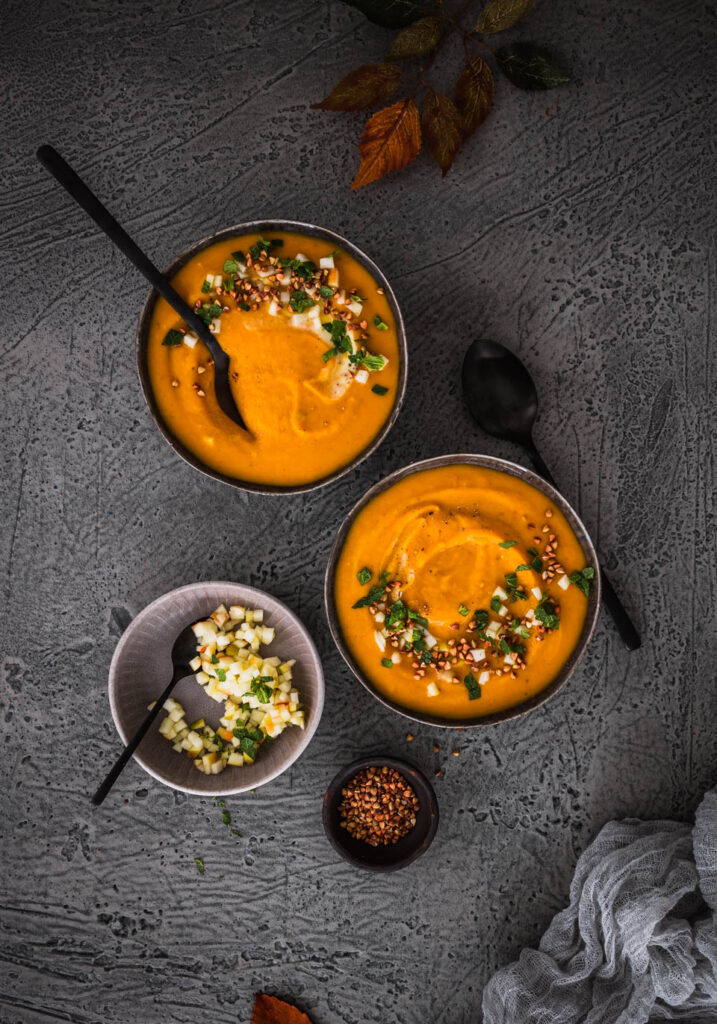 Zwei Schüssel Kürbissuppe mit schwarzen Löffeln, daneben eine Schale mit kleinen Apfelstücken.