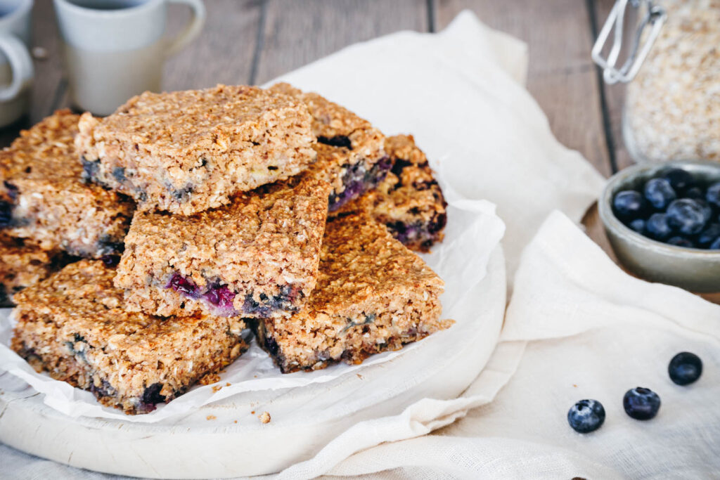 Mehrere Stücke Kuchen mit Haferflocken und Heidelbeeren auf Butterbrotpapier und weißem Holzschneidebrett.