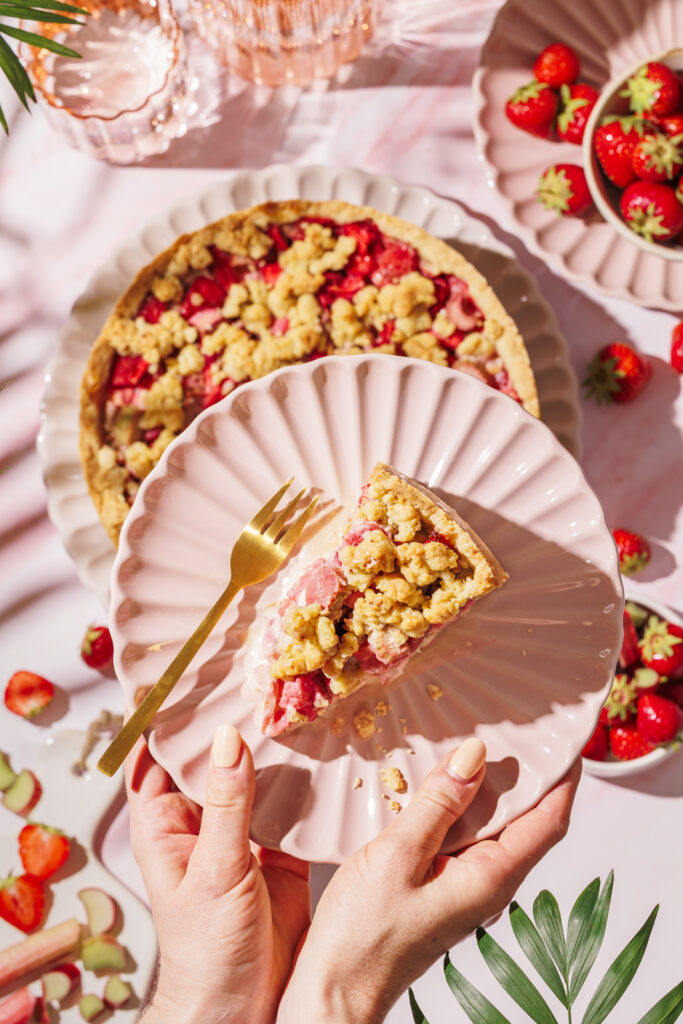 Hände halten Teller mit einem Stück Rhabarber-Streuselkuchen mit Topfencreme und Erdbeeren, daneben eine goldene Kuchengabel.