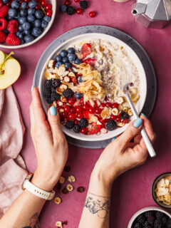 Vogelperspektive auf zwei Hände, die auf einem dunkelrosa Untergrund einen Löffel und eine Schüssel Bircher Müsli mit vielen Beeren als Topping halten.