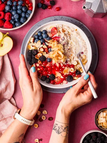 Vogelperspektive auf zwei Hände, die auf einem dunkelrosa Untergrund einen Löffel und eine Schüssel Bircher Müsli mit vielen Beeren als Topping halten.