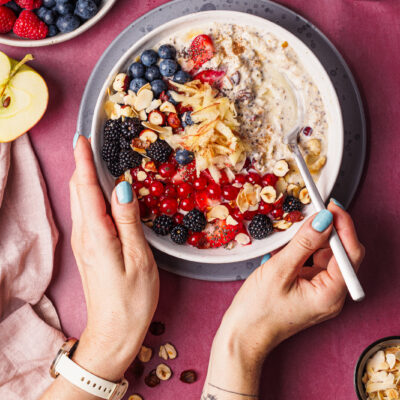Vogelperspektive auf zwei Hände, die auf einem dunkelrosa Untergrund einen Löffel und eine Schüssel Bircher Müsli mit vielen Beeren als Topping halten.