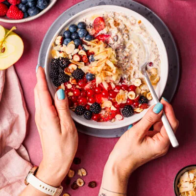 Vogelperspektive auf zwei Hände, die auf einem dunkelrosa Untergrund einen Löffel und eine Schüssel Bircher Müsli mit vielen Beeren als Topping halten.