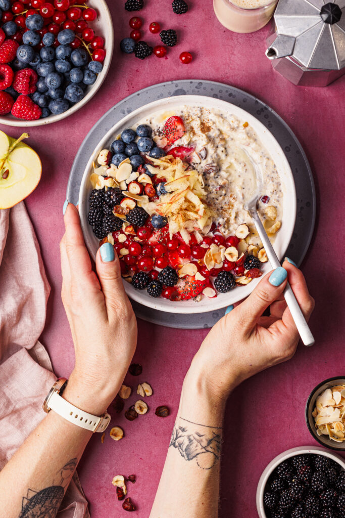 Vogelperspektive auf zwei Hände, die auf einem dunkelrosa Untergrund einen Löffel und eine Schüssel Bircher Müsli mit vielen Beeren als Topping halten.